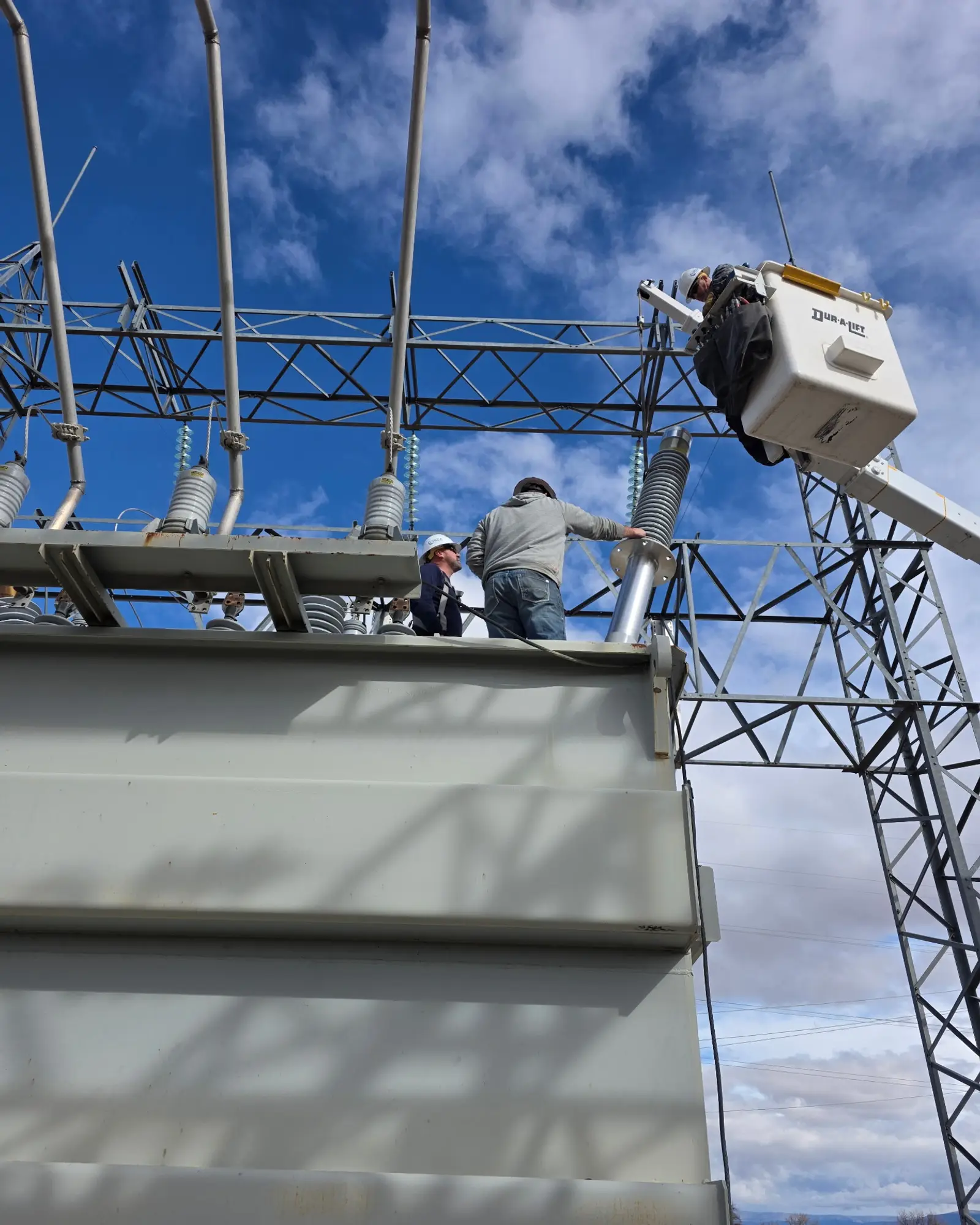 Two crew members working on top of a power transformer, with a bucket truck alongside
